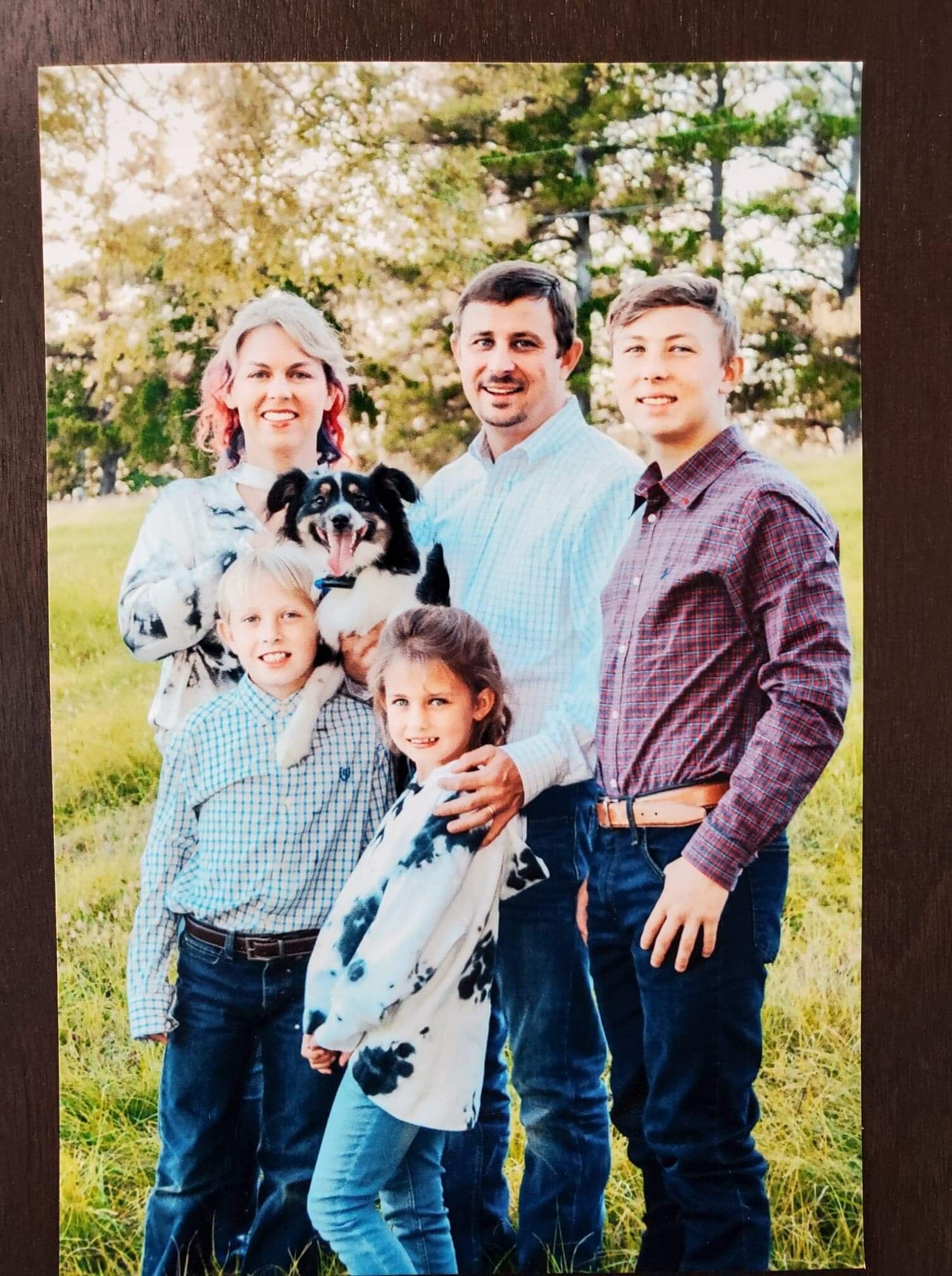 Family portrait featuring a smiling group of five people and a dog in a grassy outdoor setting. The adults and children are dressed in casual attire, with the adults in button-up shirts and the children in plaid and tie-dye. The scene captures a joyful moment in nature, emphasizing family connection and companionship.