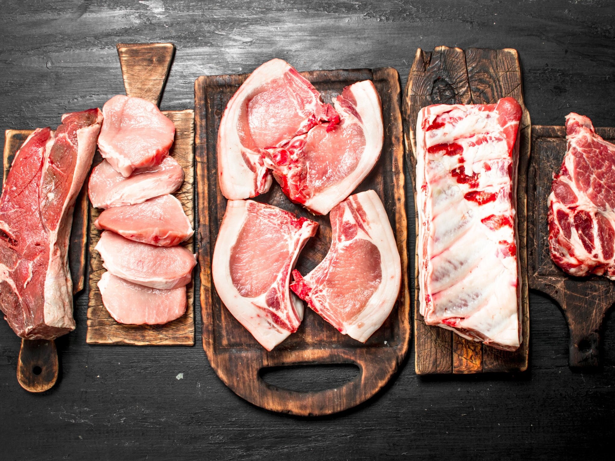 Mixed+pork+cuts Assorted raw meats displayed on wooden cutting boards, including pork chops, pork loin, and beef cuts, arranged on a dark background for culinary presentation.