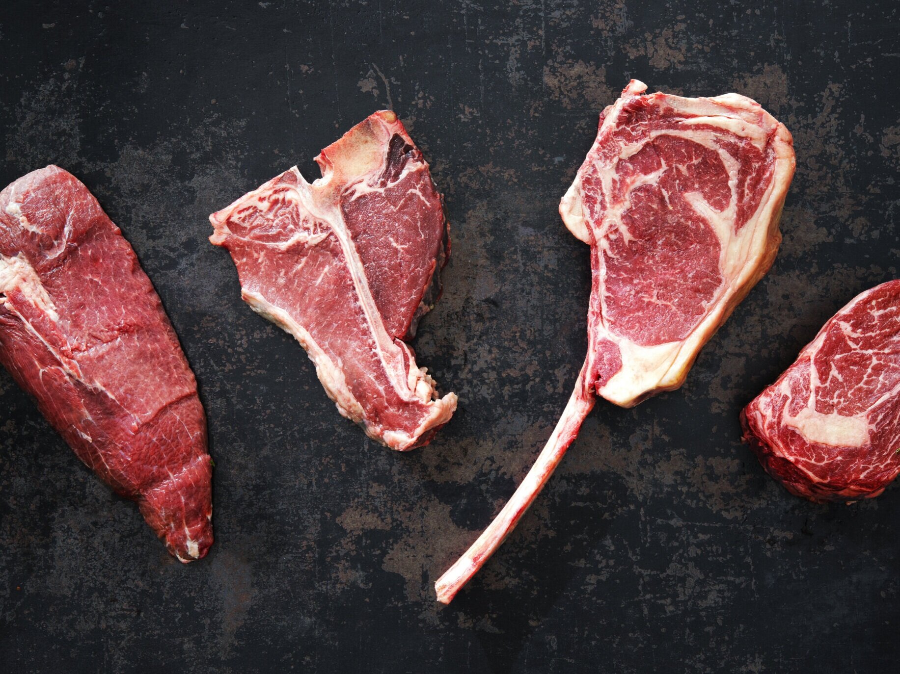 beef_porterhouse+ribeye+tenderloin+2 Assorted cuts of raw beef steak displayed on a dark surface, showcasing a variety of textures and marbling, ideal for meat lovers and culinary enthusiasts.