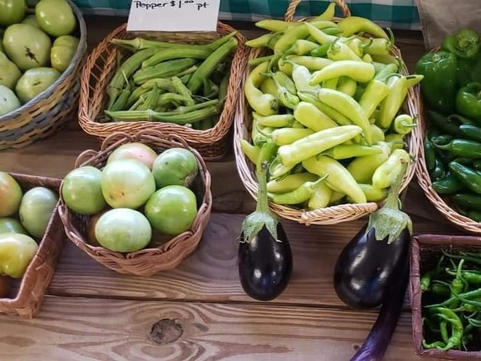 peppergreen+tomato+eggplant Fresh produce display featuring green tomatoes, okra, various types of peppers, and eggplants in woven baskets at a farmer's market.
